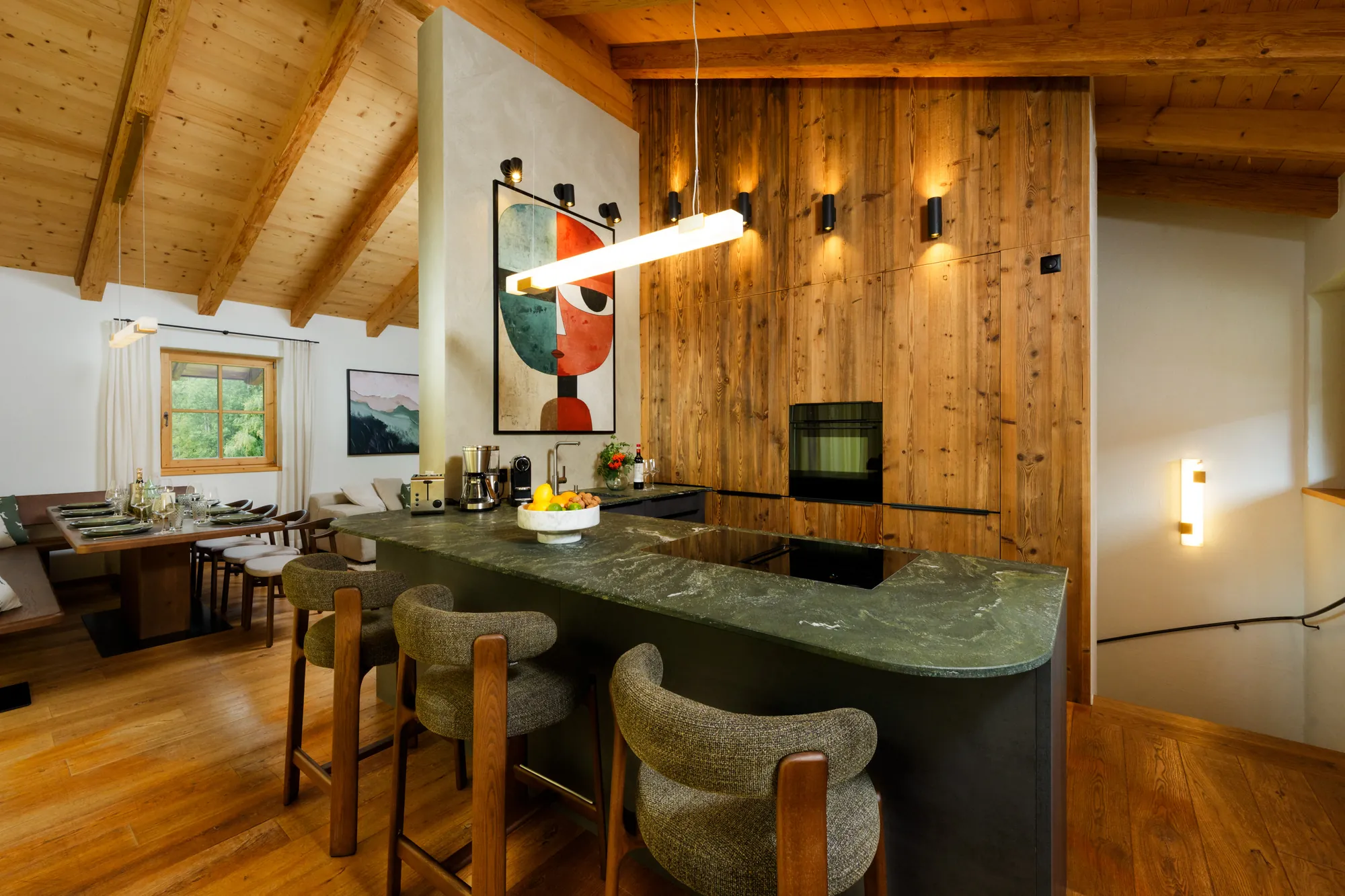 Kitchen island with green stone top and upholstered bar stools, wood-panelled wall with wall lights and dining area behind